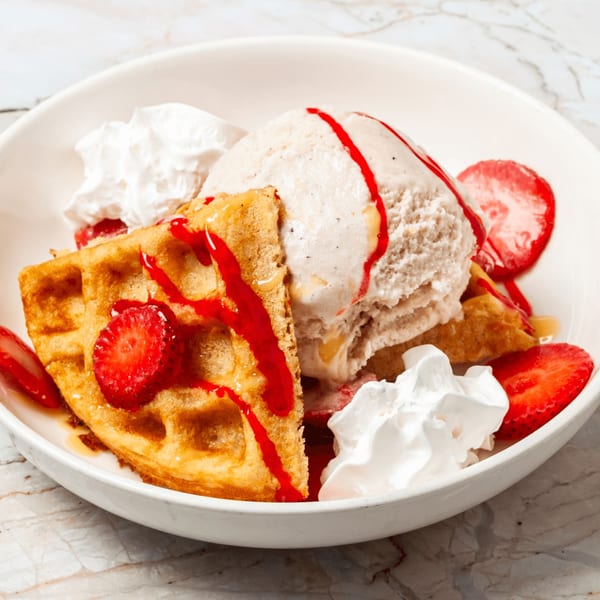 Waffle with ice cream, strawberries, whipped cream, and strawberry drizzle in a white bowl.